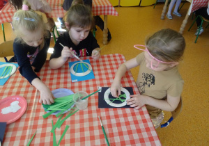 Trzy dziewczynki naklejają zielone papierowe łodygi na kartkę.